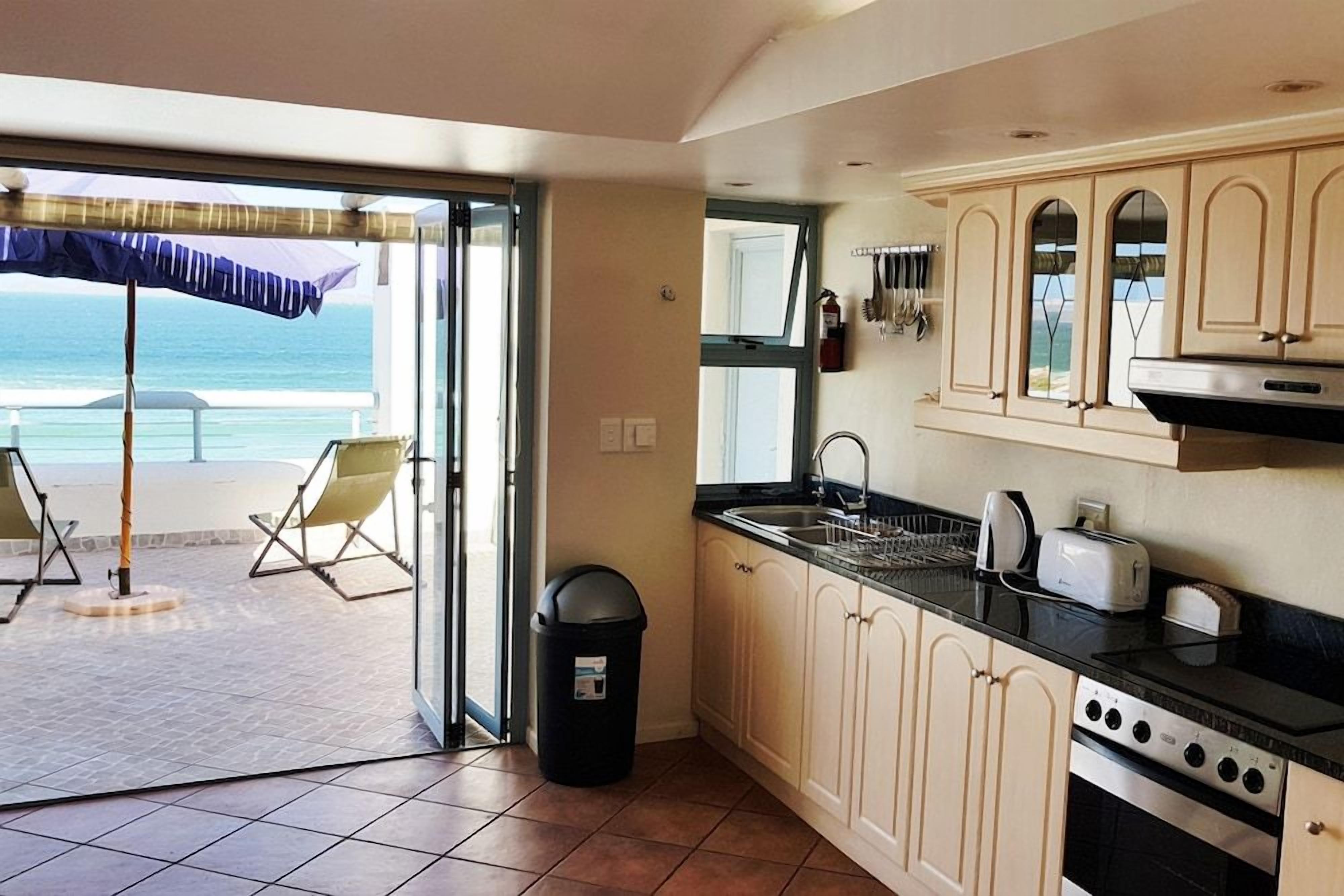 A kitchen with a view of the ocean, a trash bin, and a dining area with a chair.