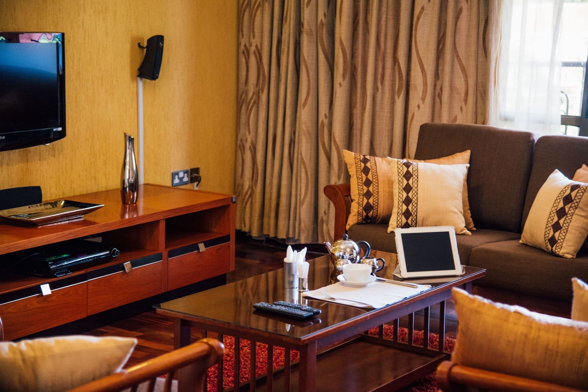 A hotel room with a flat-screen TV, a wooden coffee table with a laptop, remote controls, and a teapot.
