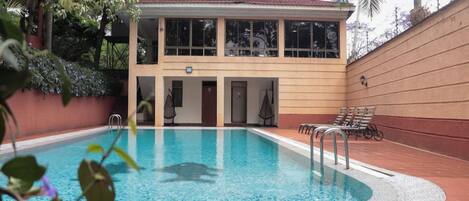 A swimming pool with a two-story building adjacent to it, featuring a covered area with seating and a door.
