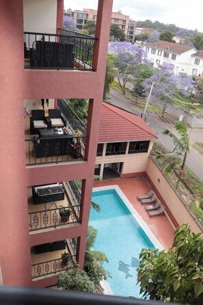 Un edificio de varios pisos con piscina en la planta baja, balcones con mobiliario exterior y vistas a casas y árboles circundantes.
