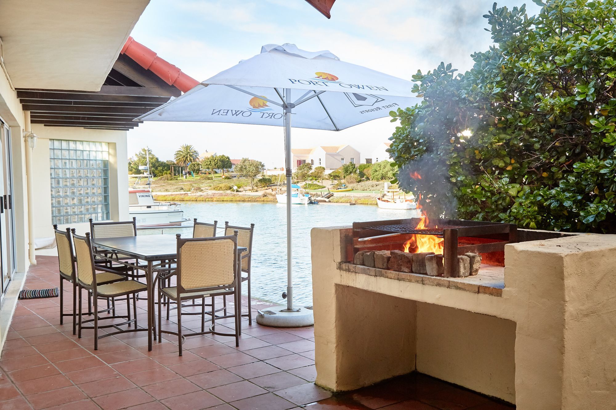 An outdoor patio with a grill, dining table, chairs, and a view of the water and boats.
