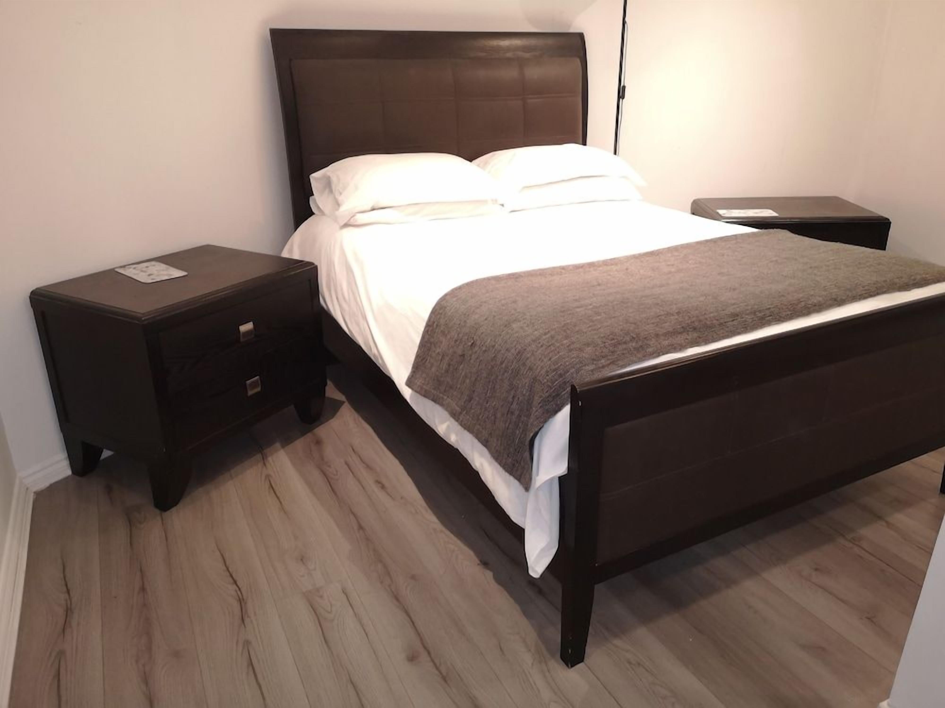 A neatly made bed with a dark headboard, a bedside table with a lamp, and a small drawer unit.