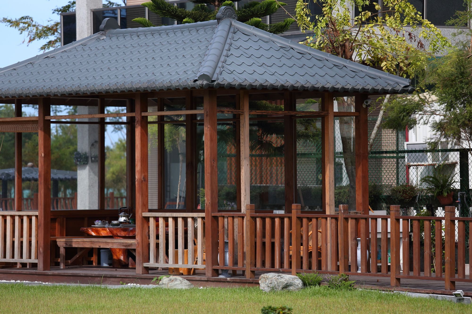 A wooden gazebo with a tiled roof, surrounded by a wooden fence, and set in a grassy area with trees in the background.