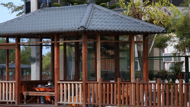 Um gazebo de madeira com telhado de azulejos, cercado por uma vedação de madeira, situado em uma área gramada com árvores ao fundo.