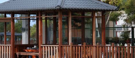 A wooden gazebo with a tiled roof, surrounded by a wooden fence, and set in a grassy area with trees in the background.