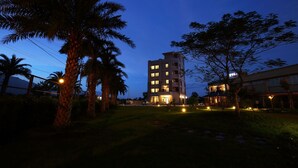 Hotel de vários andares com janelas iluminadas, cercado por palmeiras e um gramado.