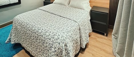 A modern bedroom with a wooden headboard, a large bed with patterned bedding, a nightstand, a mirror, and wall-mounted artwork.