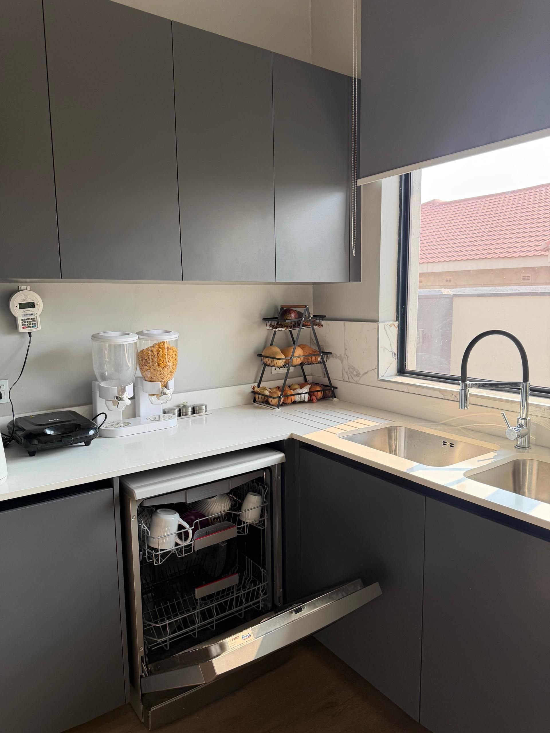 A modern kitchen with grey cabinets, a built-in dishwasher, a coffee machine, and a window with a view of a red-tiled roof.