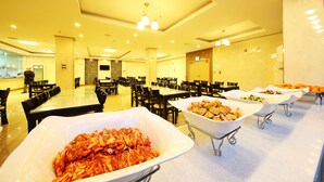 Zona de dinning d'un hotel amb diverses taules i cadires, una barra de buffet amb diferents plats, i una persona al fons.