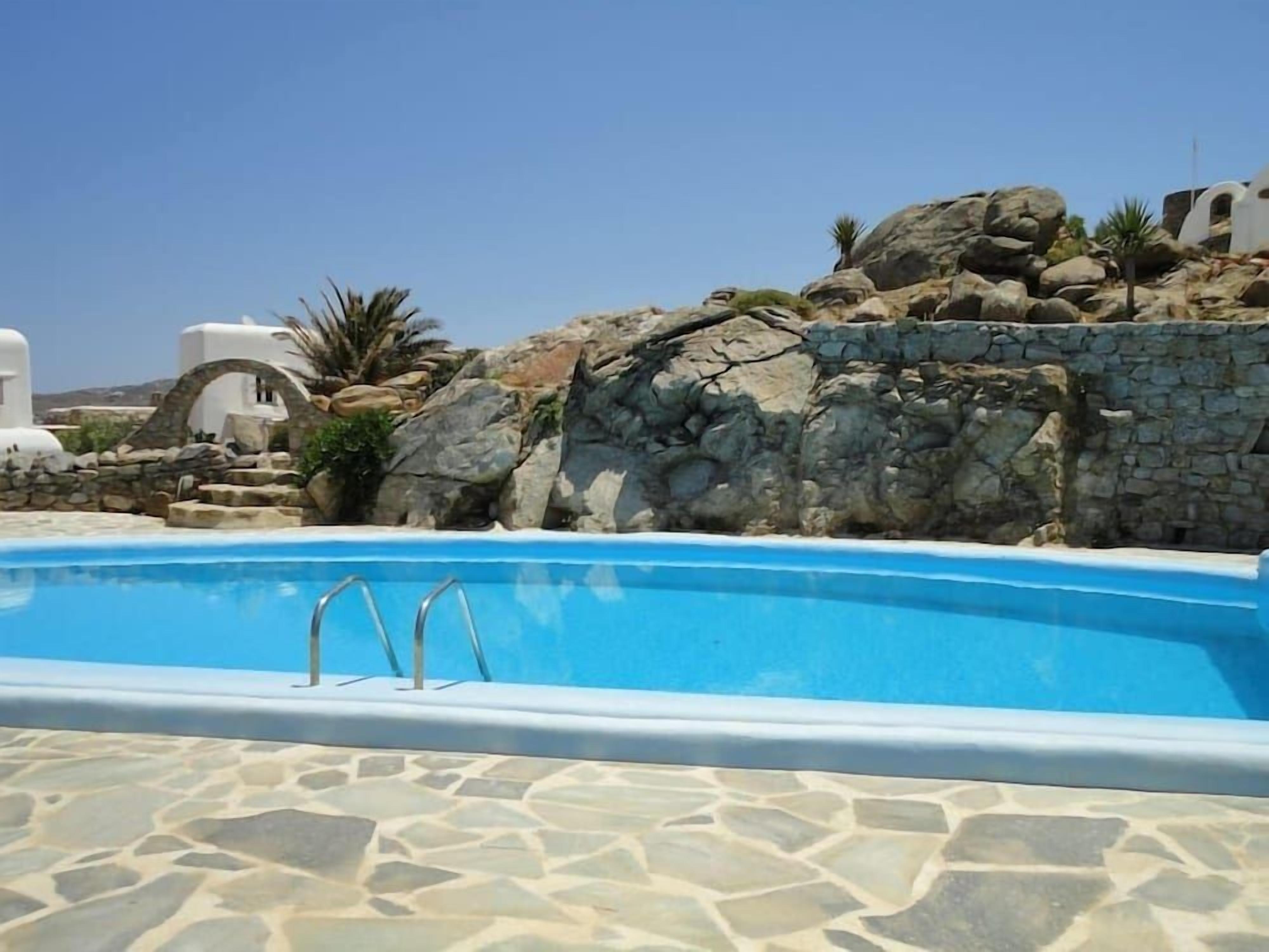 A swimming pool with stone tiles, surrounded by white buildings and rocky terrain.