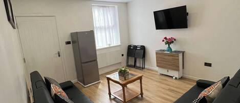 Una sala de estar con dos sofás negros, una mesa de centro, un televisor en la pared, un refrigerador y una ventana con cortinas.