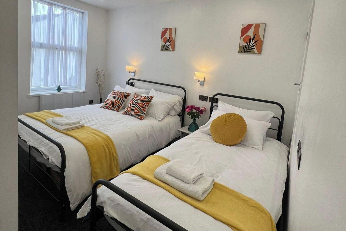 Two single beds with white bedding and yellow accents, a window with sheer curtains, and wall-mounted lamps.