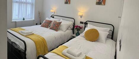 Dos camas individuales con ropa de cama blanca y detalles en amarillo, una ventana con cortinas translúcidas y lámparas fijadas a la pared.