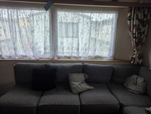 A room with a sofa set against a wall, a window with sheer curtains, and a patterned curtain on the right. - 3 Bedroom Seaview Cabin Sleeps 8 - Balcony & Pool (Skegness)