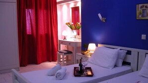 A hotel room with a bed, a bedside table with a bottle and glass, a chair, a desk with a lamp, and a red curtain. - Terpsichori Hotel (Chania)