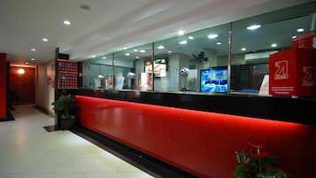 Uma área de recepção com balcão vermelho, divisórias de vidro e uma tela digital exibindo conteúdo.