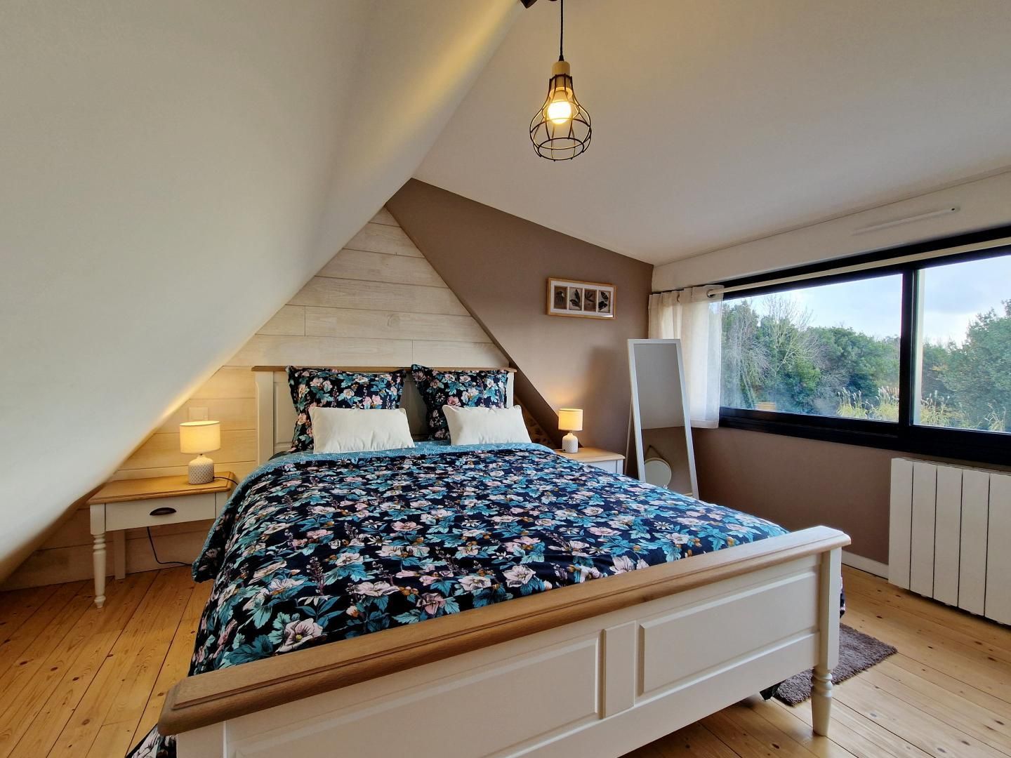 A bedroom with a sloped ceiling, a bed with floral bedding, bedside lamps, a window with a view, and a hanging light fixture.