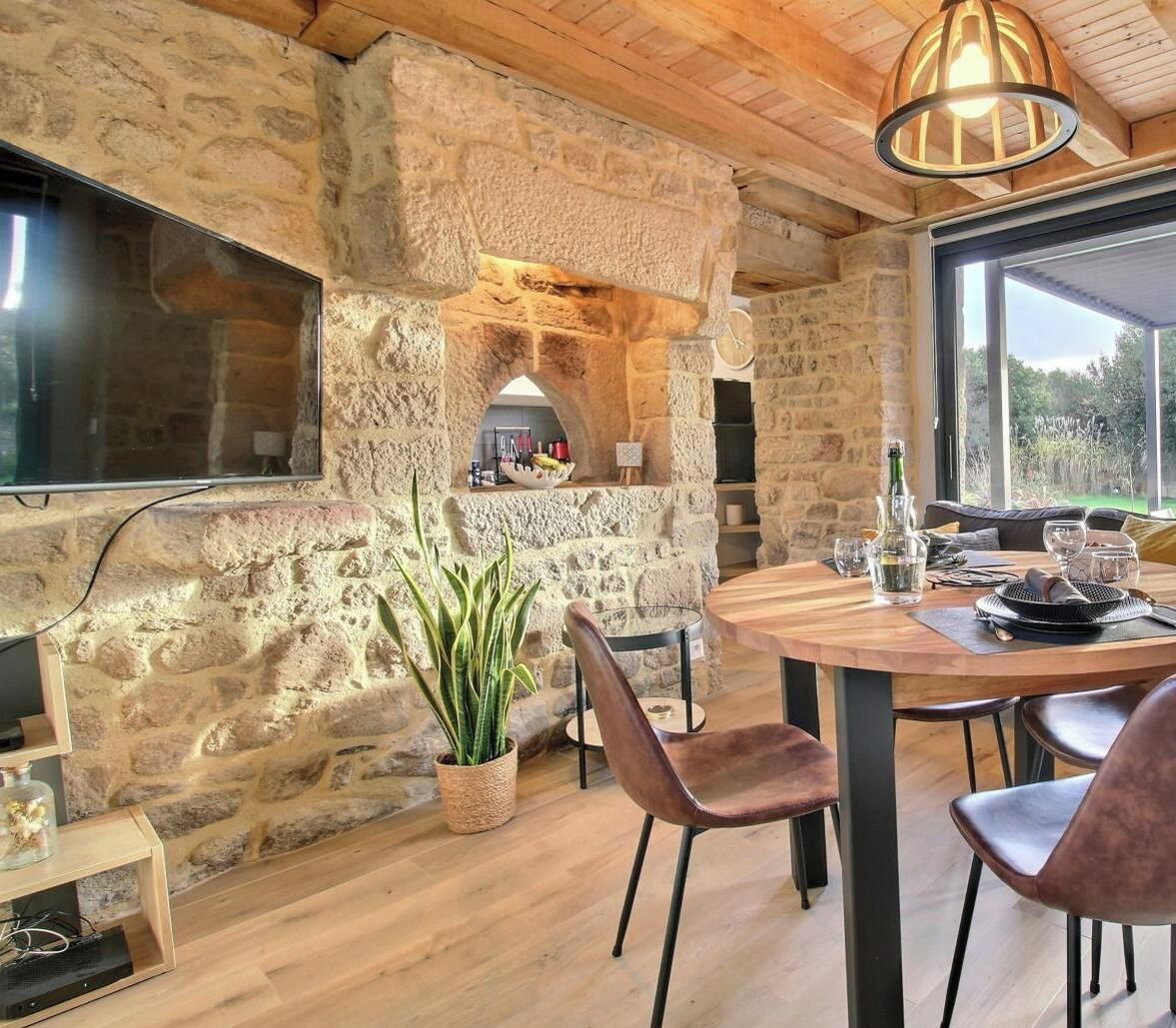 Une pièce aux murs de pierre, dotée d’une cheminée, d’une table à manger avec des chaises et d’une grande fenêtre.
