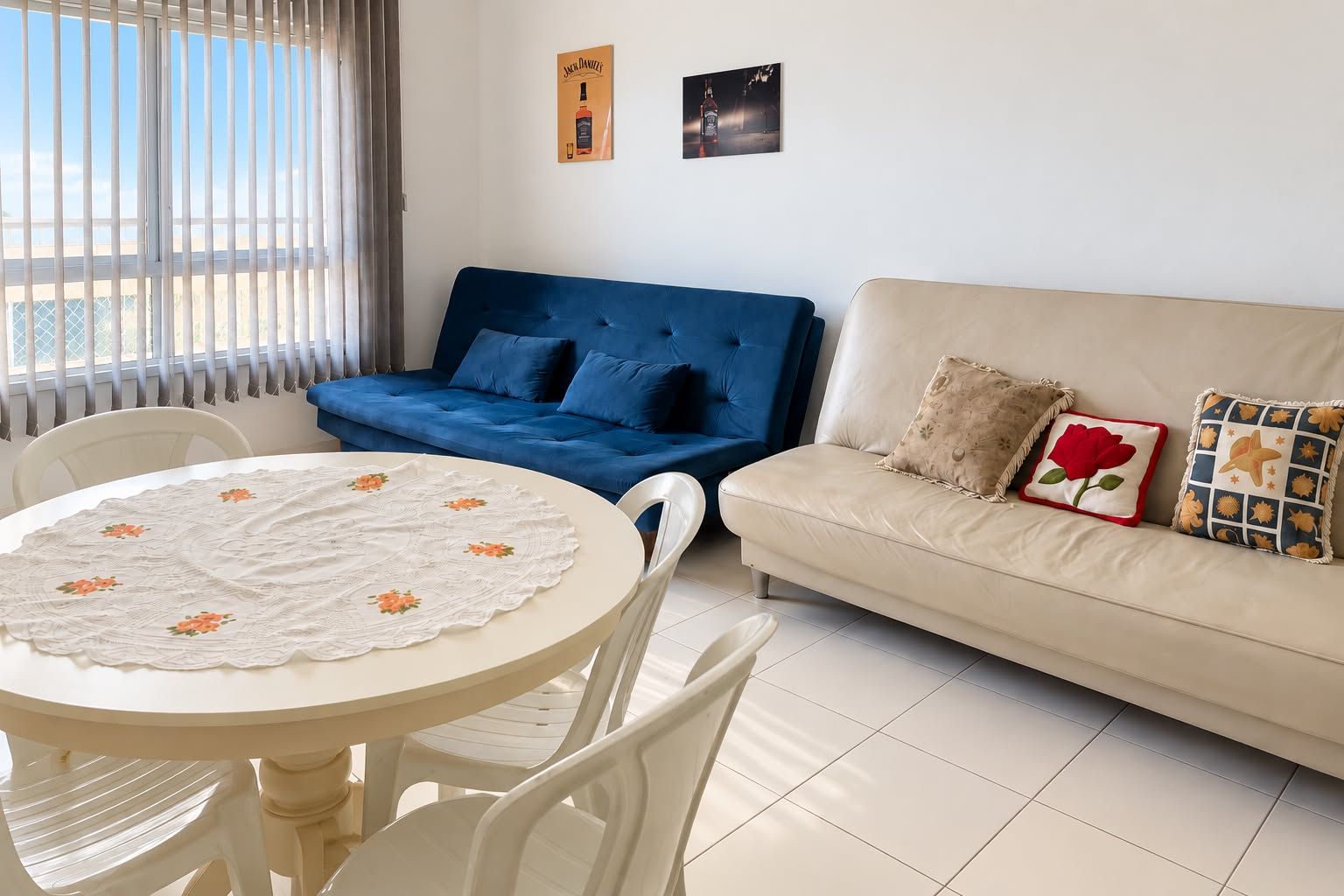 Een woonkamer met een blauwe bank, een beige bank, een ronde tafel met bloemen tafelkleed en twee stoelen.