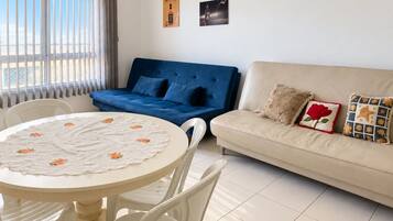 Een woonkamer met een blauwe bank, een beige bank, een ronde tafel met bloemen tafelkleed en twee stoelen.
