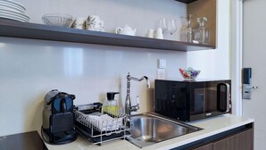 A modern kitchen with a stainless steel sink, a coffee maker, a dish drying rack, a microwave, and a shelf with glassware and dishes. - Hôtel Au Picardy (Nice)