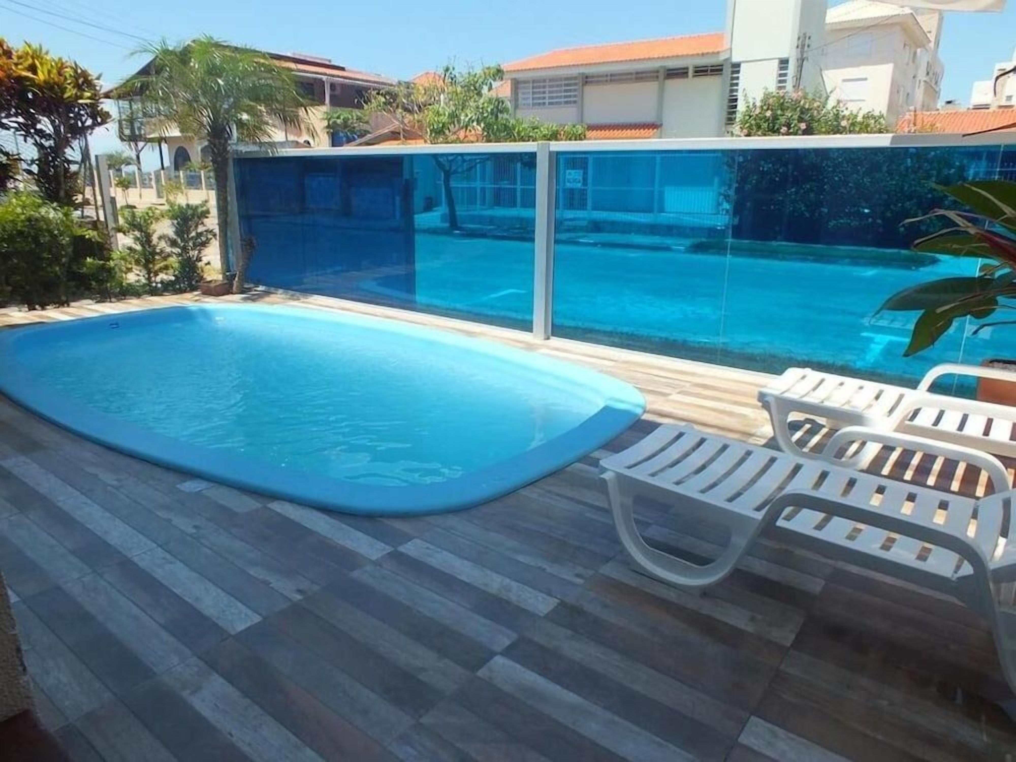 Una piscina con superficie de agua azul cristalina, rodeada por una terraza de madera y sillas de plástico blanco.