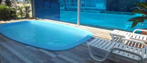 Una piscina con superficie de agua azul cristalina, rodeada por una terraza de madera y sillas de plástico blanco.