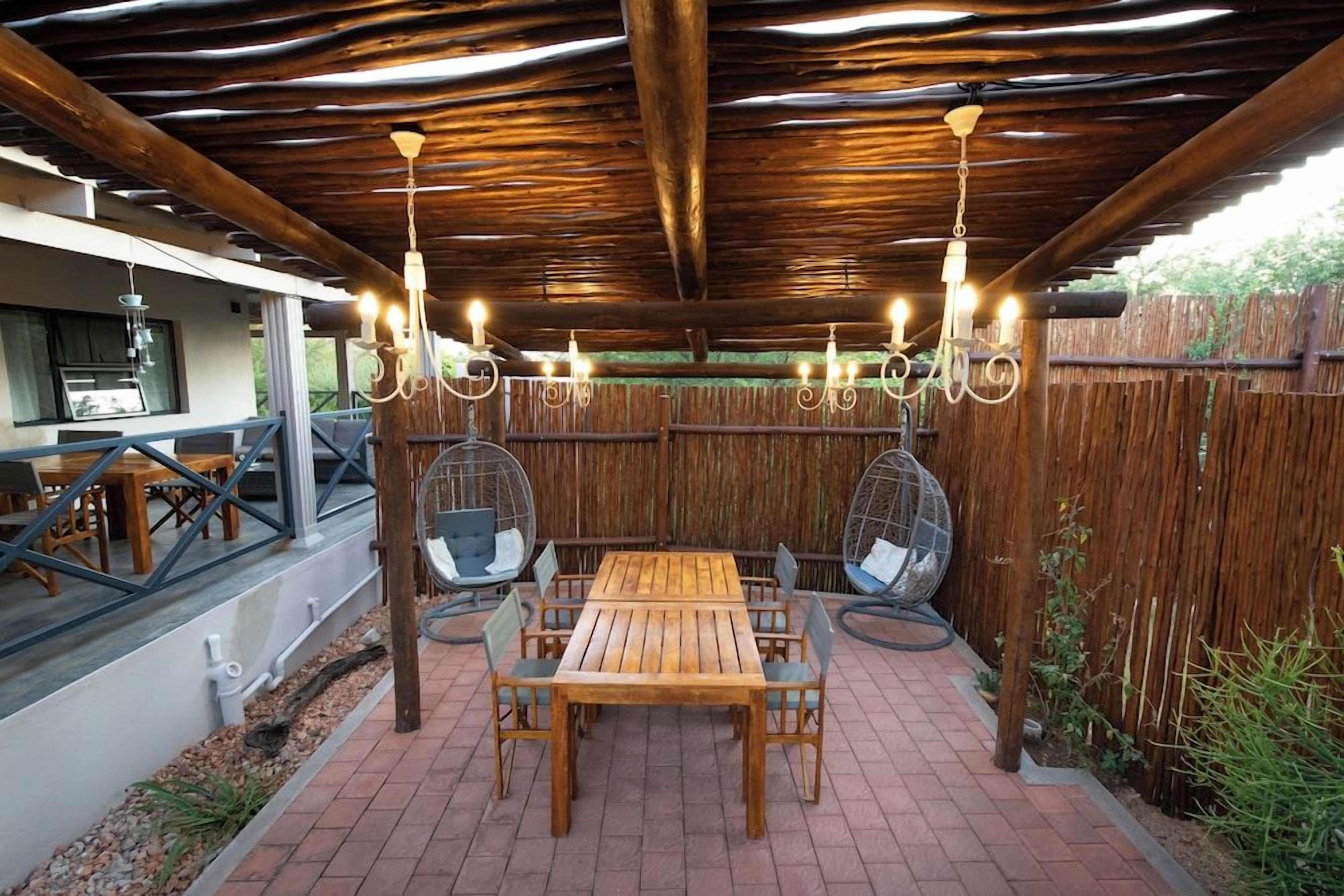 Eine Terrasse im Freien mit einem Holztisch und -stühlen, umgeben von einem Bambuszaun, unter einer Holzpergola mit hängenden Lichtern.