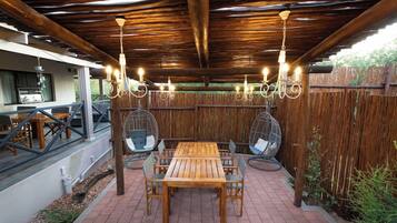 Eine Terrasse im Freien mit einem Holztisch und -stühlen, umgeben von einem Bambuszaun, unter einer Holzpergola mit hängenden Lichtern.