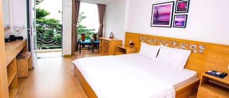 A hotel room with a large bed, a desk, a chair, and a balcony with a view of greenery.