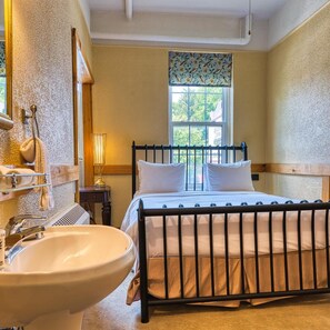 A bedroom with a bed, a window, a bedside table, a lamp, and a bathroom with a sink and mirror.