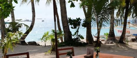 A wooden table and chairs set on a sandy beach with a beach umbrella, surrounded by palm trees and a view of the ocean.