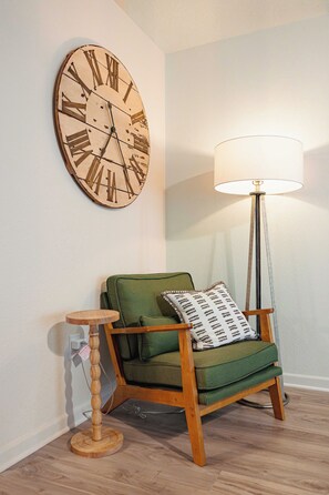 Een kamer met een houten vloer, een groene fauteuil met een kussen met dessin, een rond houten bijzettafeltje, een vloerlamp en een grote wandklok.