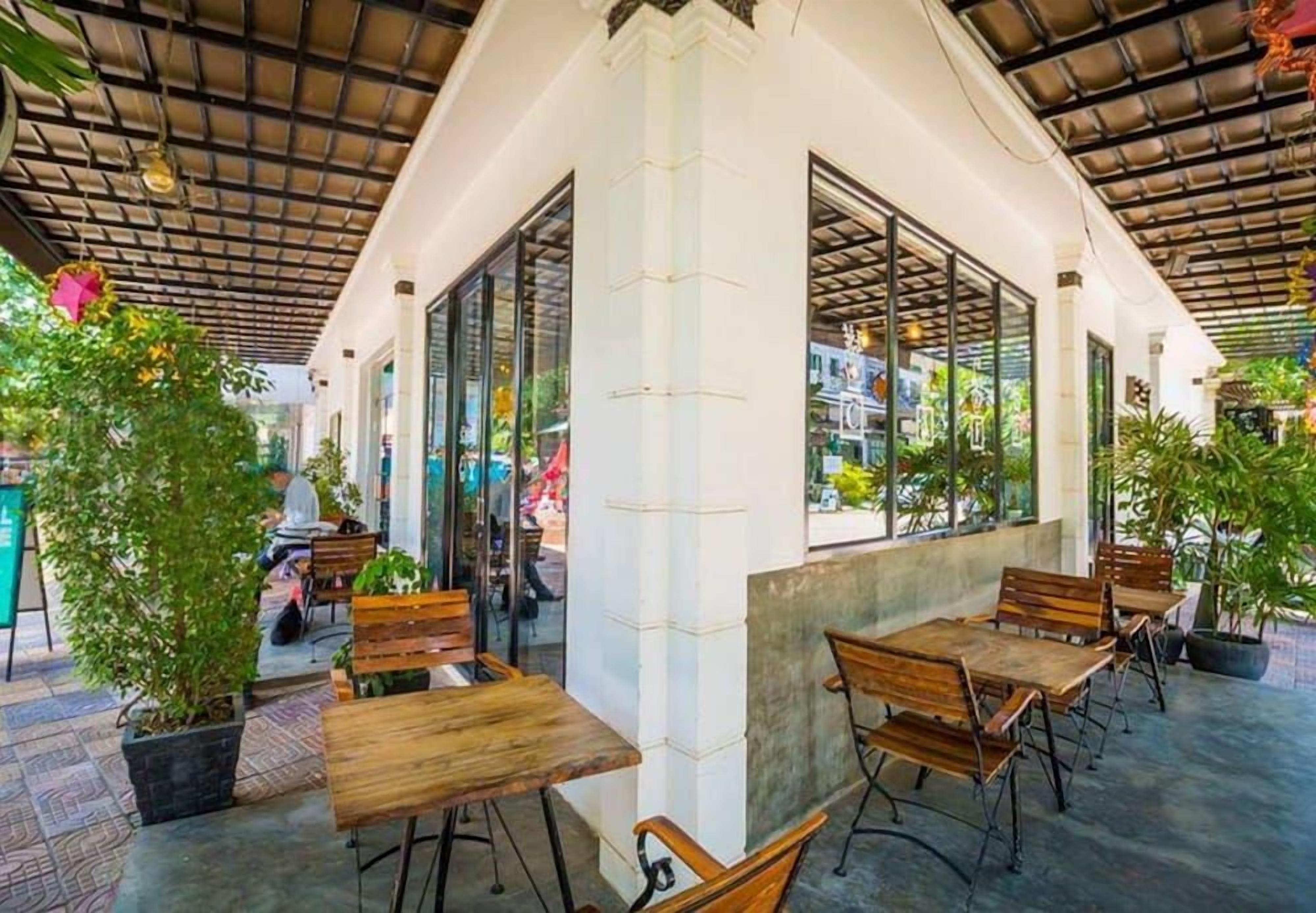 An outdoor cafe area with wooden tables and chairs, potted plants, and large windows.