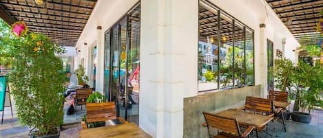 An outdoor cafe area with wooden tables and chairs, potted plants, and large windows.