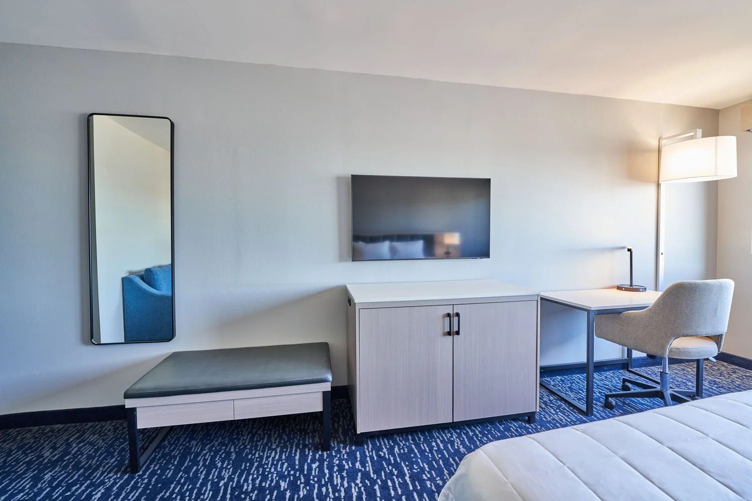 Ein Hotelzimmer mit einem Bett, einem Schreibtisch, einem Sessel, einer Bank, einem Schrank, einem Spiegel und einer Wandleuchte.