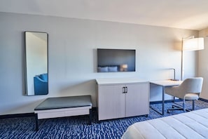 A hotel room with a bed, a desk, a chair, a bench, a cabinet, a mirror, and a wall-mounted lamp. - King Bed Harbor View (Fiarhaven)