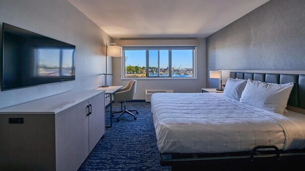 A hotel room with a bed, desk, chair, TV, and a window with a view. - King Bed Harbor View (Fiarhaven)