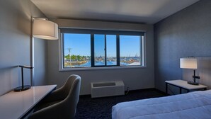 A hotel room with a large window, a bed, a desk, a chair, and a bedside table with a lamp. - King Bed Harbor View (Fiarhaven)