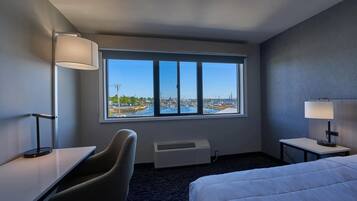 Quarto de hotel com uma grande janela, cama, escrivaninha, cadeira e mesa de cabeceira com abajur.