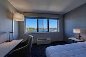 Habitación de hotel con un ventanal grande, una cama, un escritorio, una silla y una mesita de noche con lámpara.
