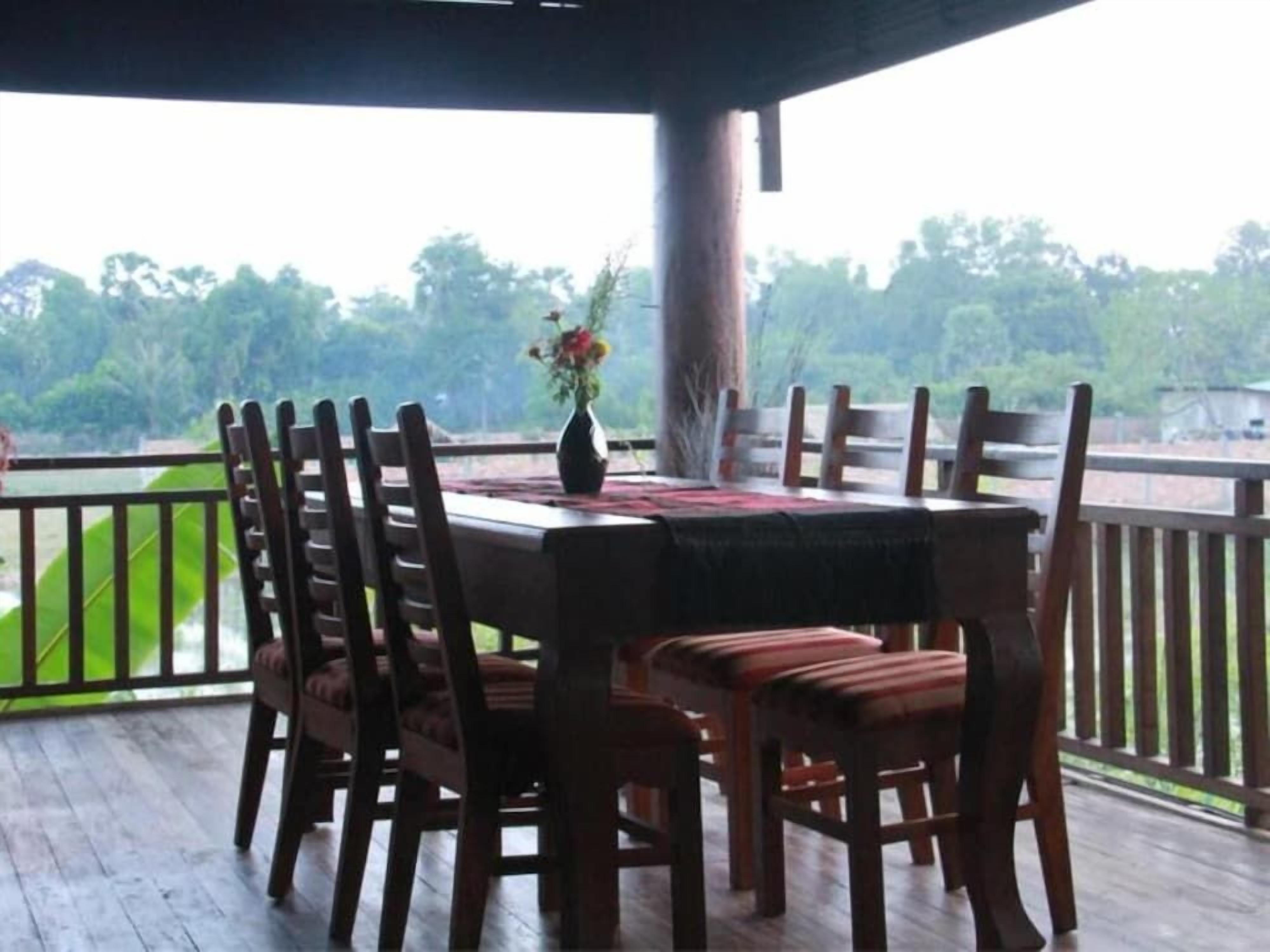 A covered outdoor dining area with a table and chairs, a vase with flowers, and a view of greenery.