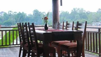A covered outdoor dining area with a table and chairs, a vase with flowers, and a view of greenery.