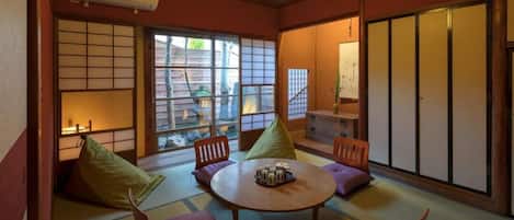 A traditional Japanese room with tatami flooring, a low table with cushions, sliding doors, and a view of the outside.