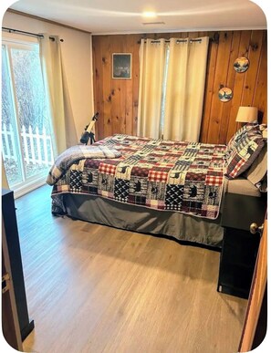 A bedroom with a wooden floor, a bed with a quilt, a nightstand, a lamp, and a window with curtains. - Cozy Ski Cabin w/ Fenced Yard 10 Min to Mt Snow, 20 Min to Stratton,Dog Friendly (Wardsboro)