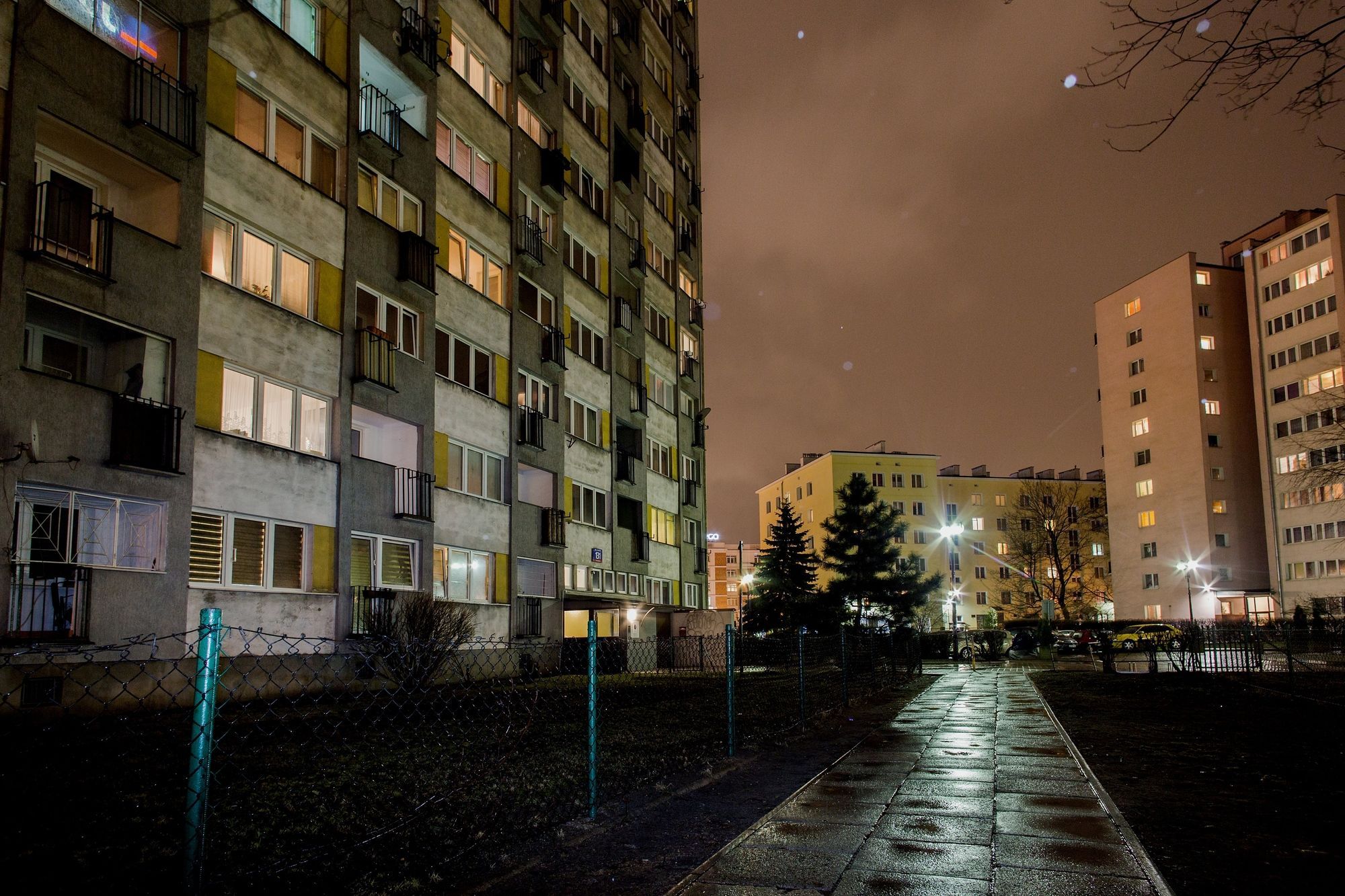 Nattebillede af en høj bygning med lejligheder og altaner, samt en våd fortov.