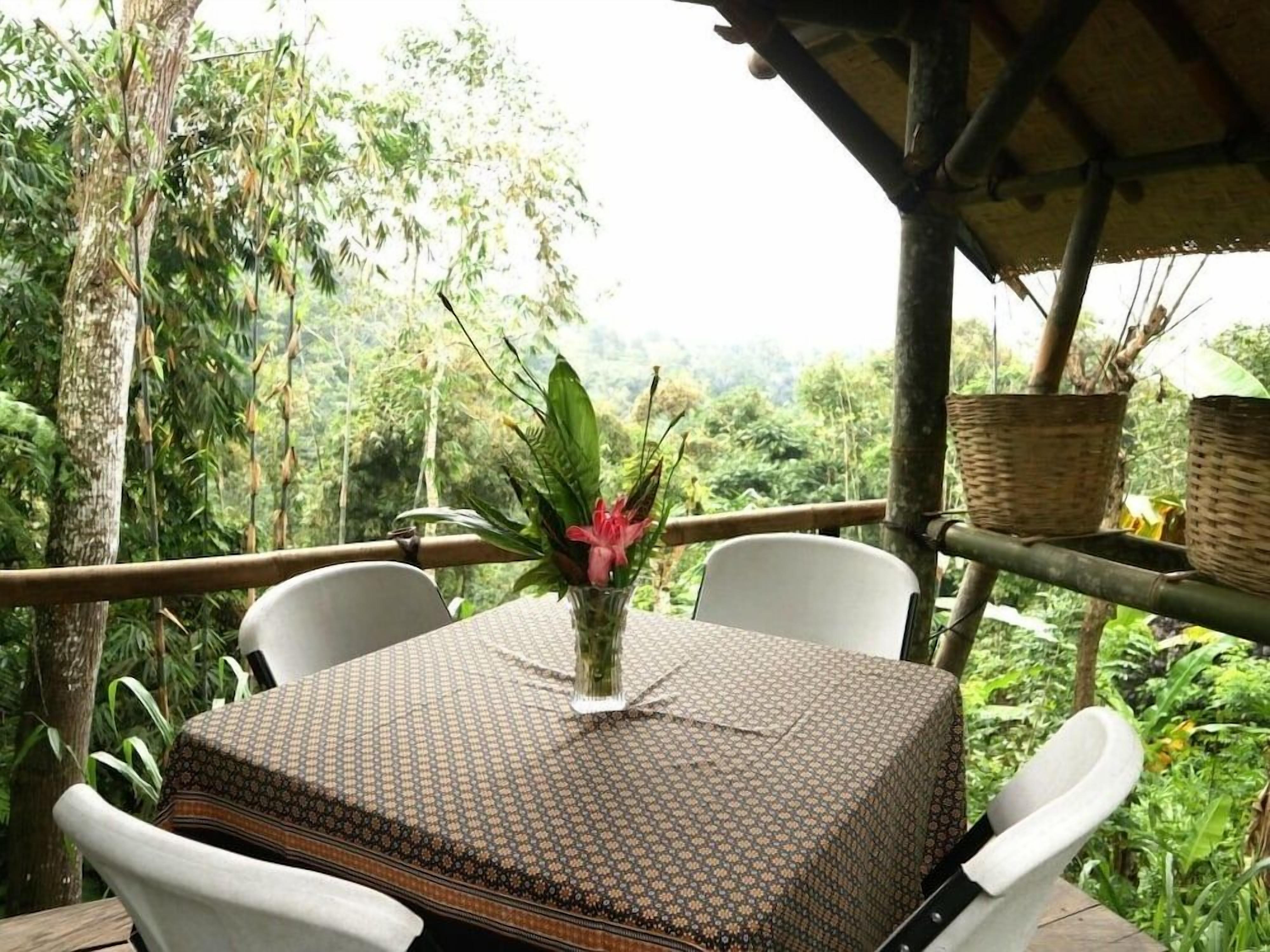 An outdoor seating area with a table covered by a patterned tablecloth, surrounded by white chairs, and a vase with a flower.