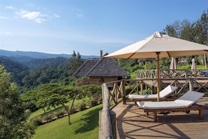 A wooden deck with lounge chairs and a large umbrella, overlooking a lush valley with a thatched-roof building.