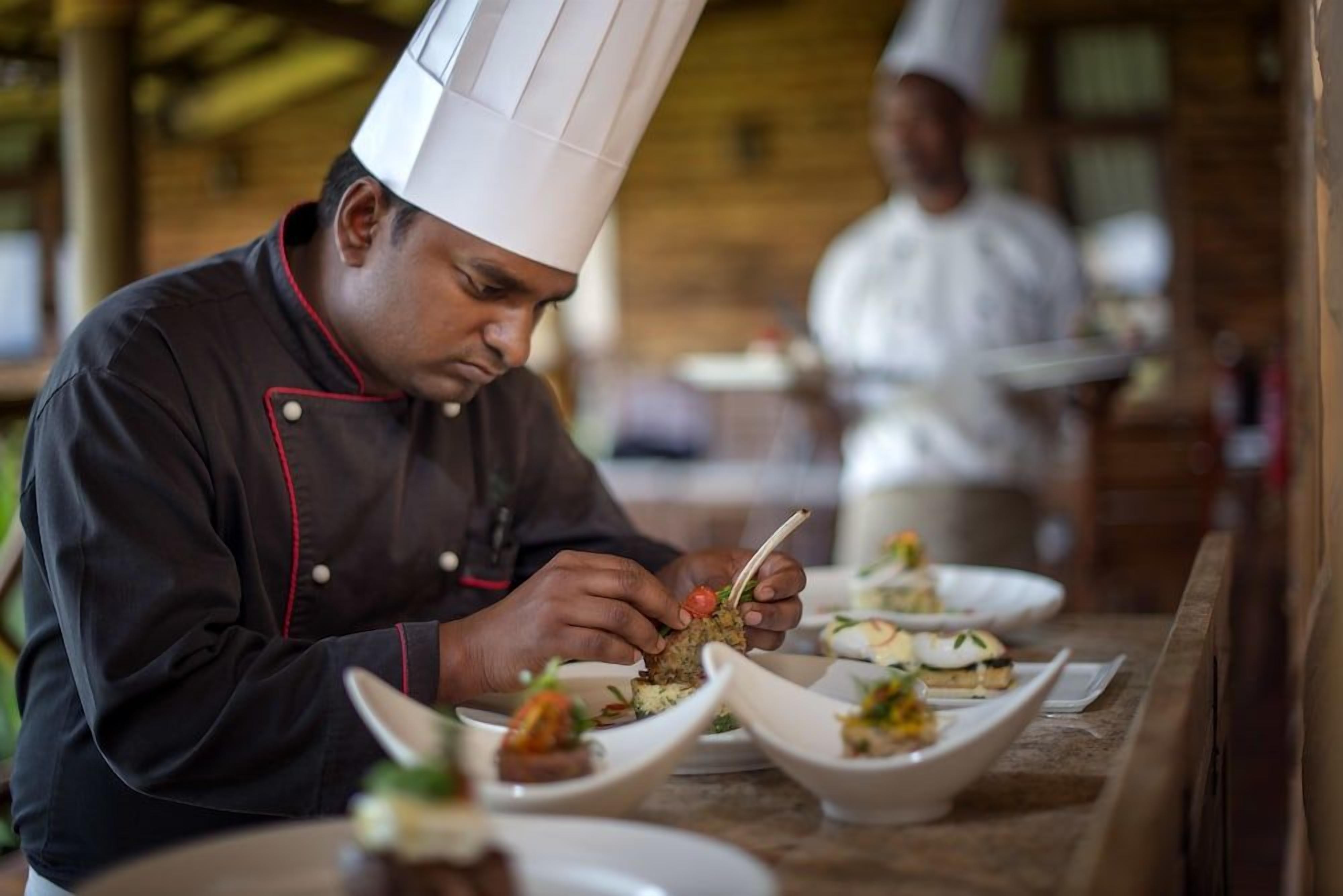 A chef in a black uniform with red trim, preparing food at a counter with white plates.