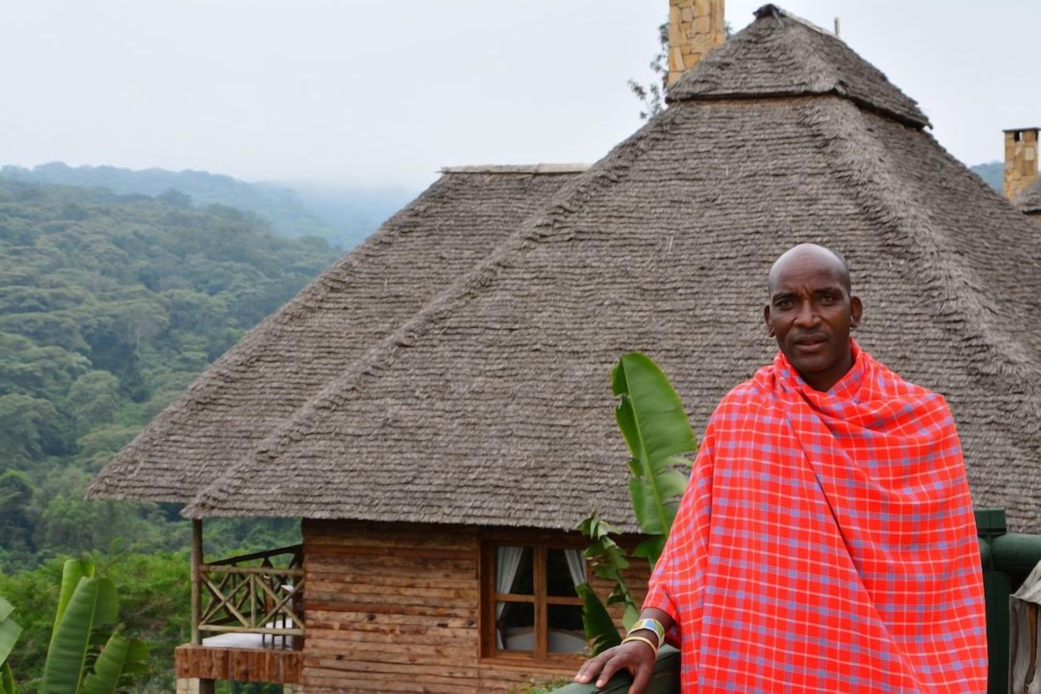 A person standing in front of a thatched-roof hut with a wooden structure and a view of a forested landscape.